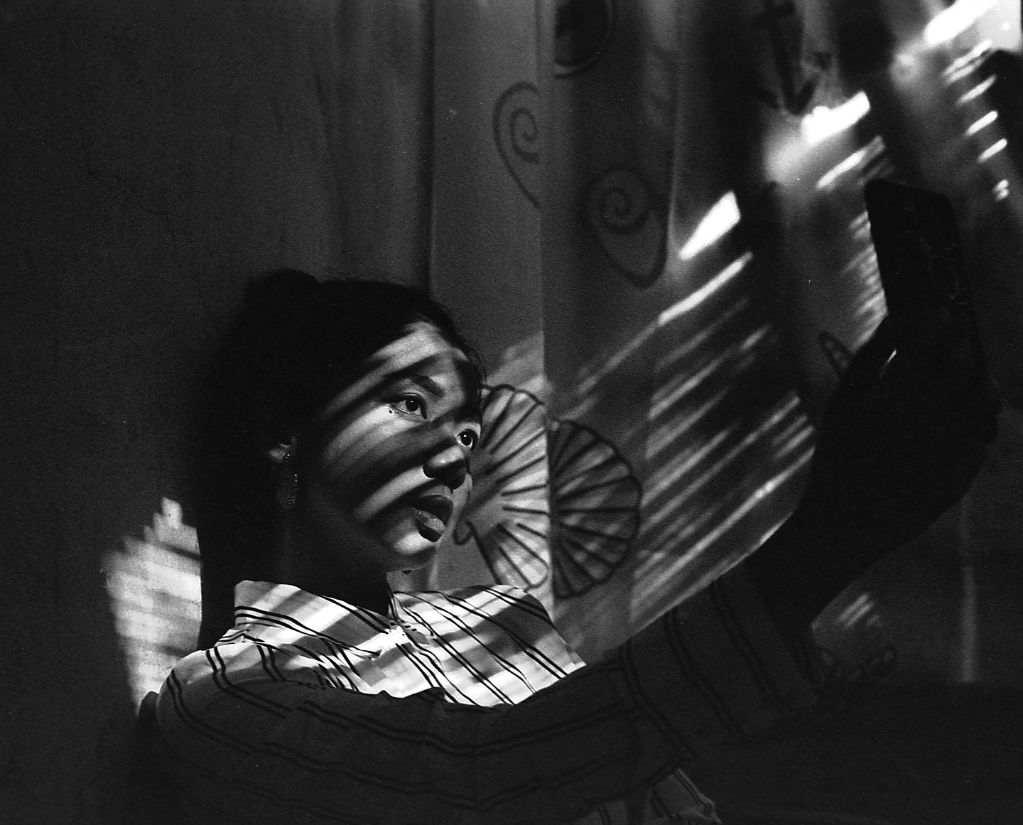 A woman in a striped shirt is taking a selfie, with shadows falling across her face and a patterned curtain behind her.