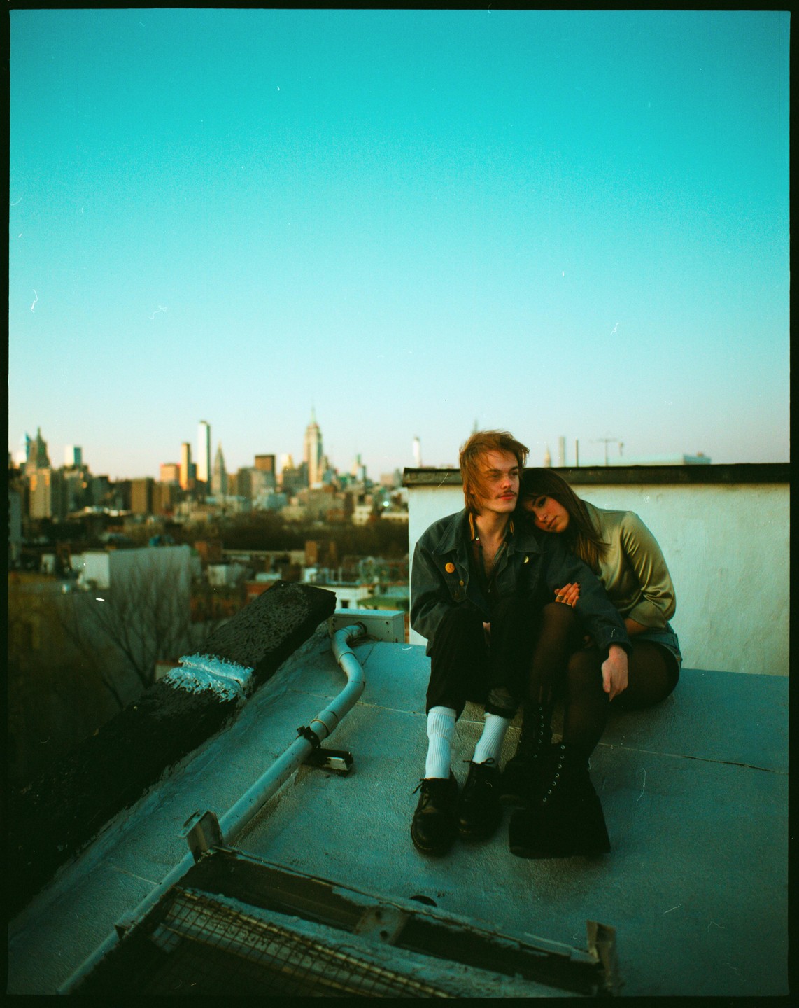 A couple sits on a rooftop overlooking a city skyline at dusk.