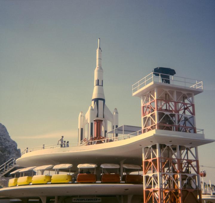 A large white rocket stands next to a building with a people mover ride and a tall tower.