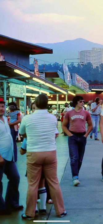 A crowd of people walk along a street with buildings and mountains in the background.
