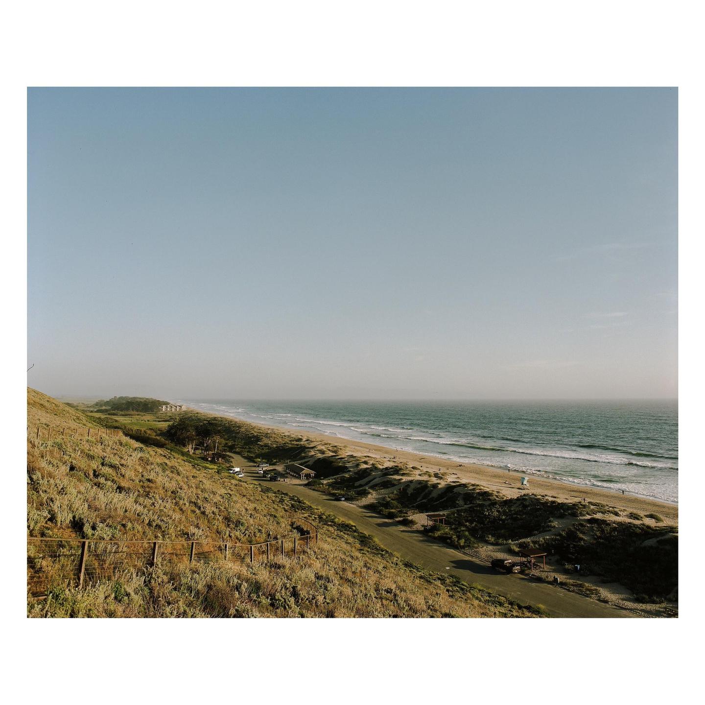 A long stretch of beach and ocean is visible from a grassy hillside.