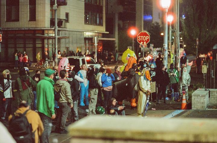 A crowd of people in costumes and masks gather on a city street at night, with a stop sign and buildings in the background.