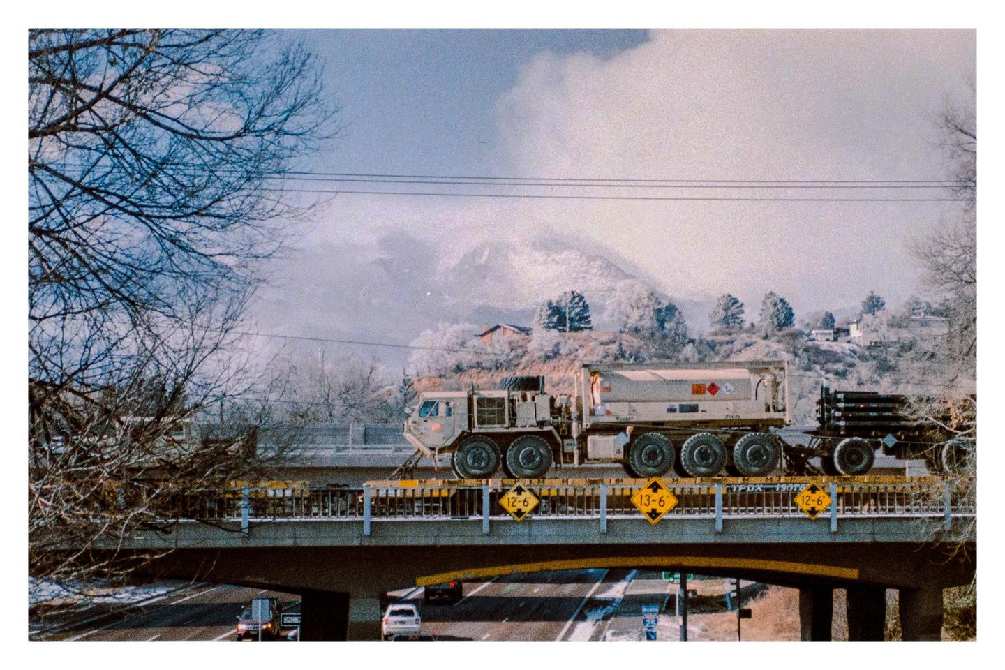 A large truck with a tanker trailer drives across a bridge over a highway, with snow-covered mountains and trees in the background.