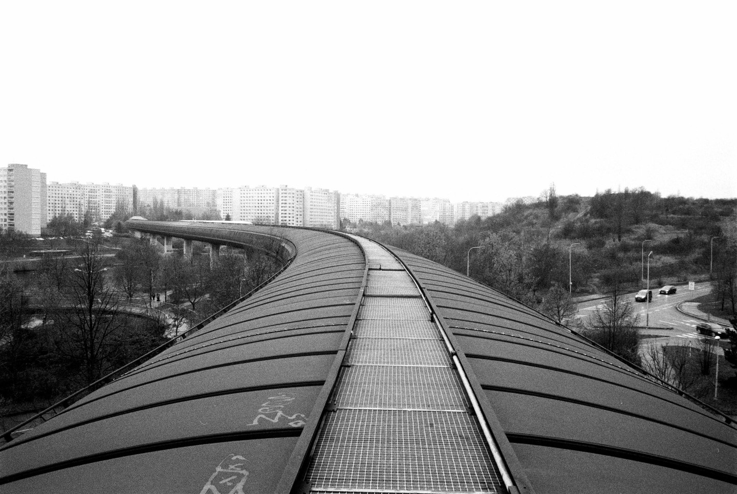 A long, curved pedestrian bridge with a metal grate walkway extends over a road and park toward a city skyline.