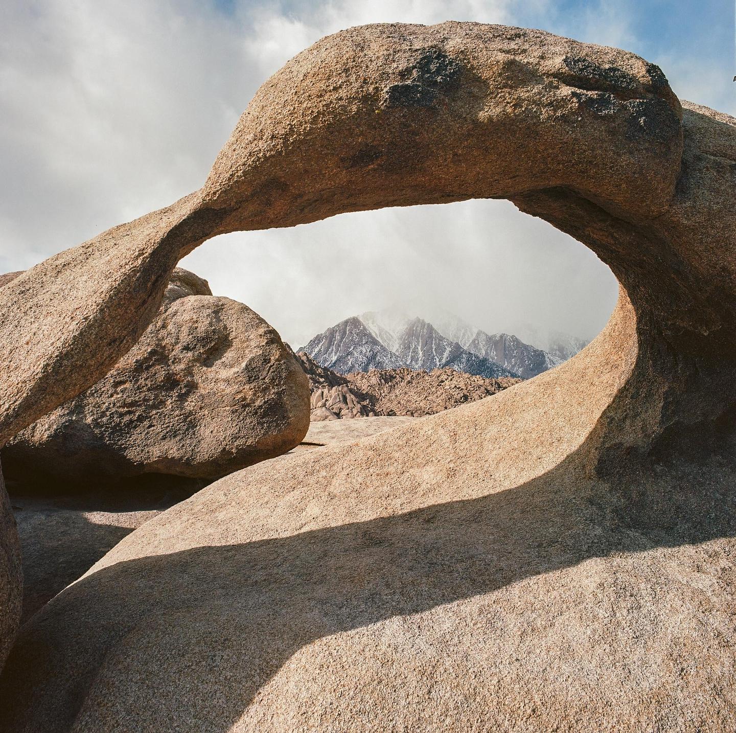 A natural rock arch frames snow-capped mountains under a cloudy sky.