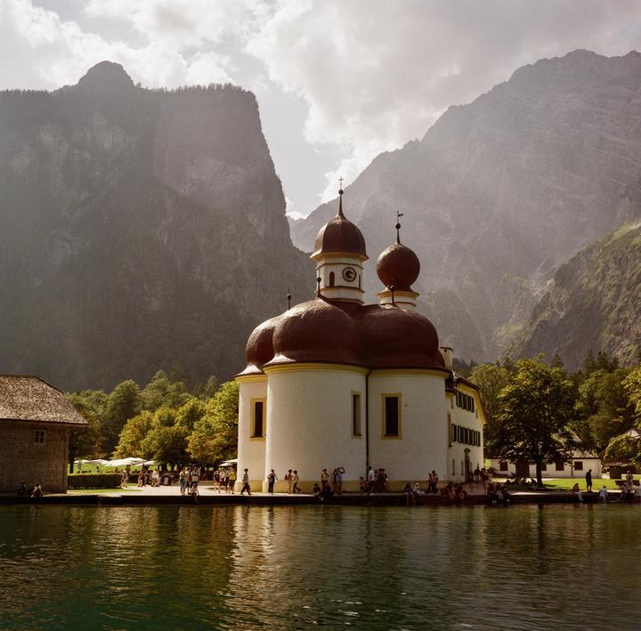 A white church with red domed roofs stands on the edge of a lake, with mountains rising in the background.