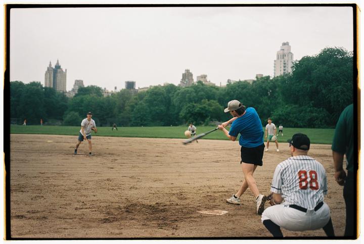 A man swings a baseball bat at a ball on a dirt field with buildings and trees in the background.