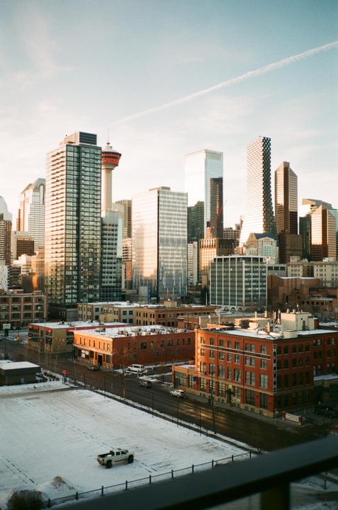 A cityscape of downtown Calgary shows tall buildings and a tower under a bright sky, with snow-covered ground and streets in the foreground.