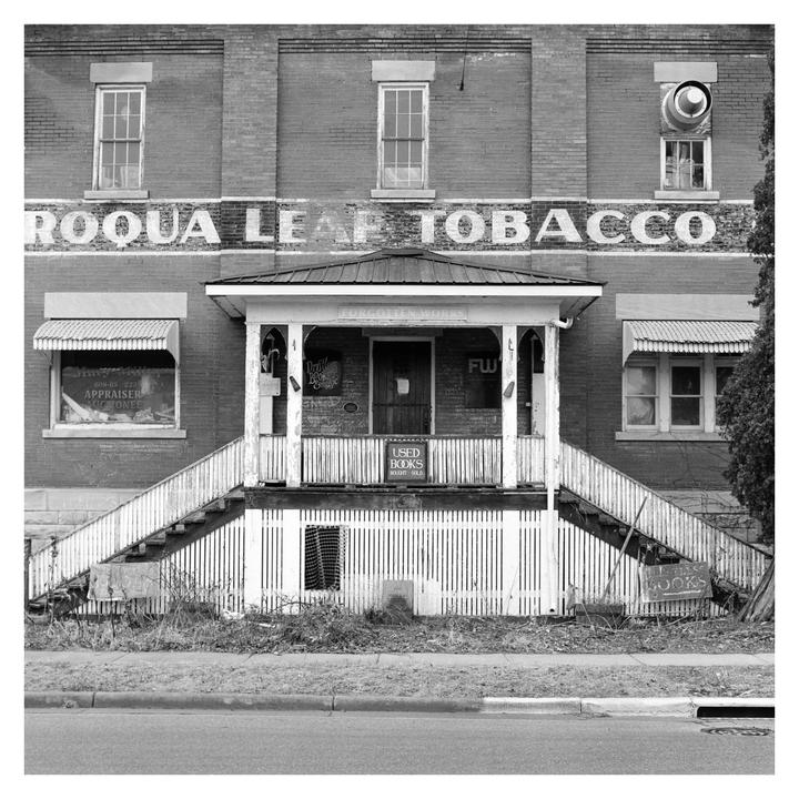 A brick building with a porch and two staircases has "ROQUA LEAF TOBACCO" painted on its facade.