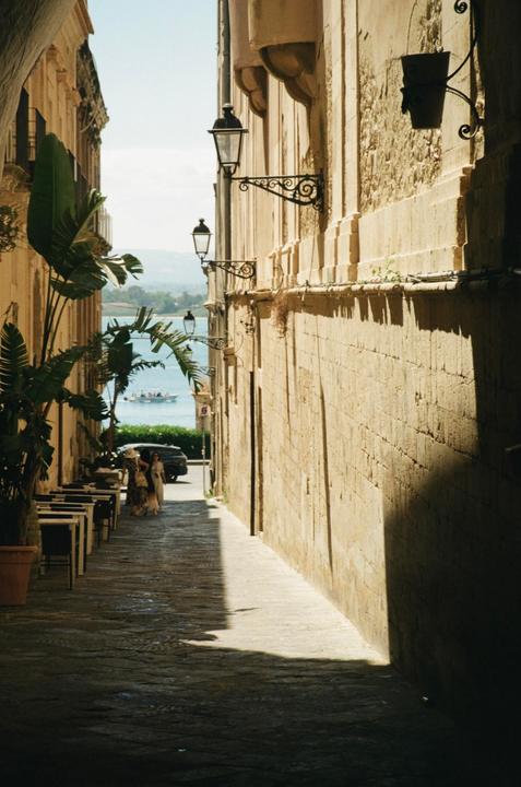 A narrow street with stone buildings and streetlights leads to a body of water with a boat.