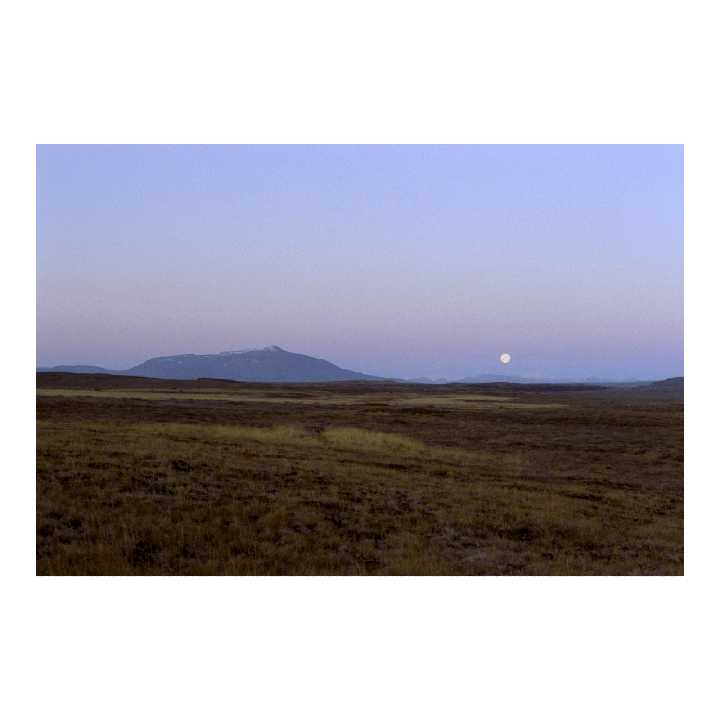 A full moon hangs over a vast field with mountains in the distance at dusk.
