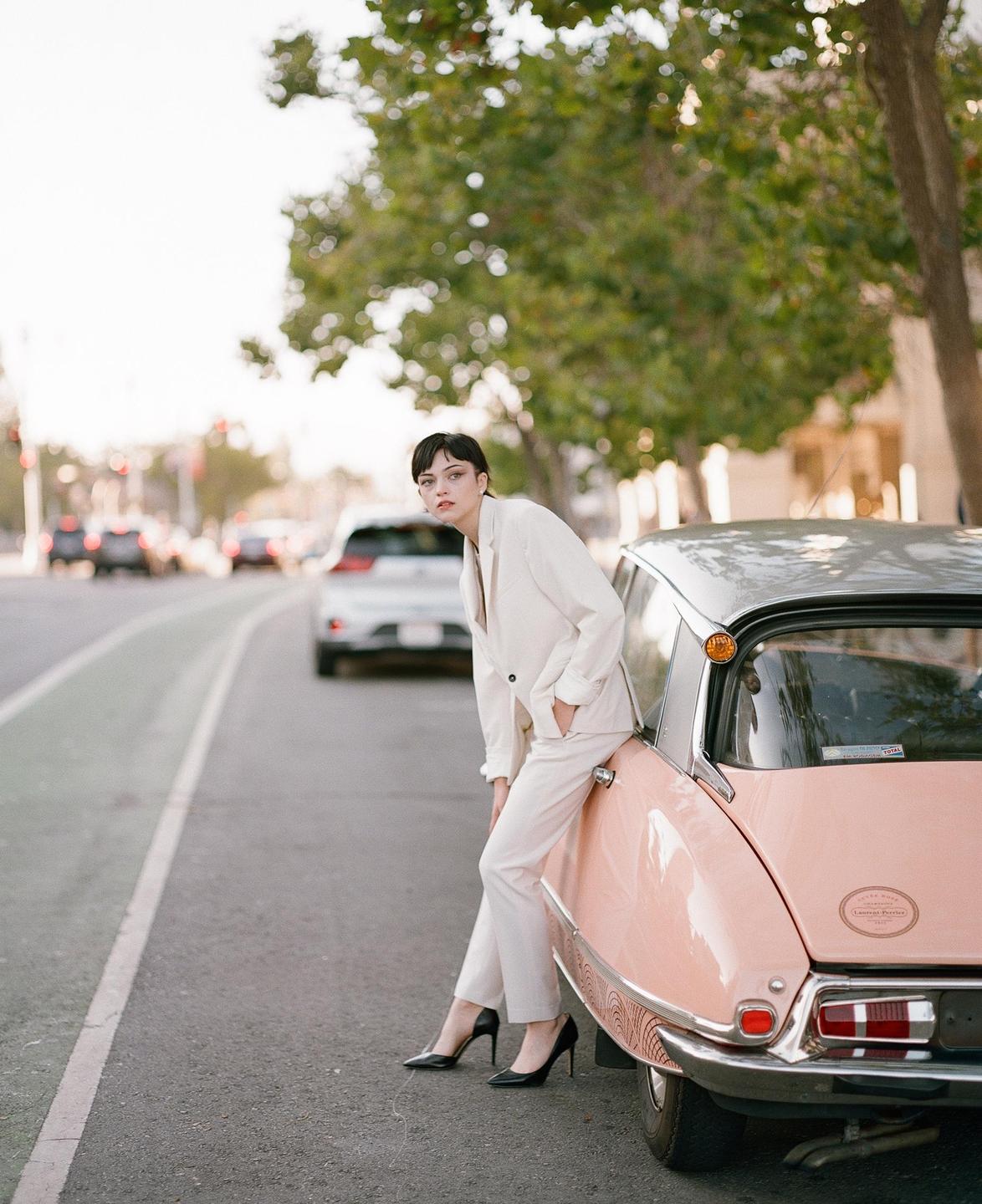 A woman in a white suit leans against a pink vintage car on a street.
