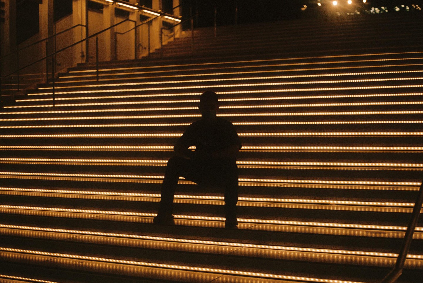 A man sits silhouetted on a wide staircase with illuminated steps at night.