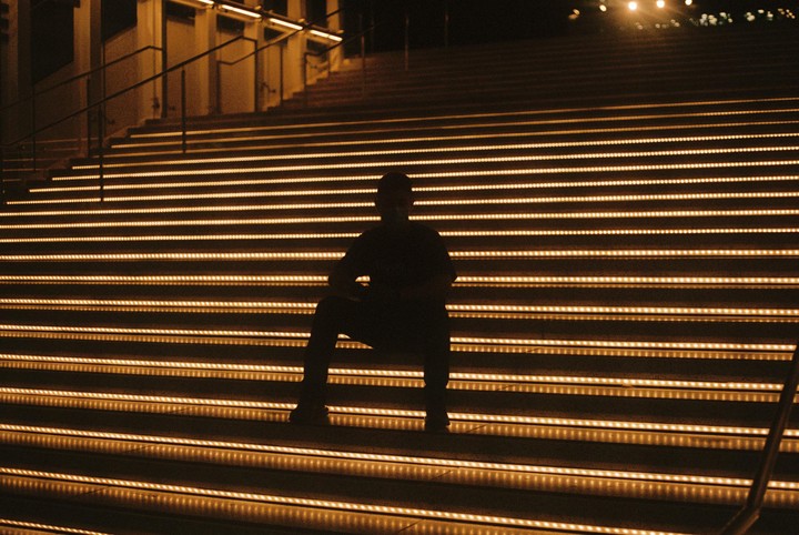 A man sits silhouetted on a wide staircase with illuminated steps at night.