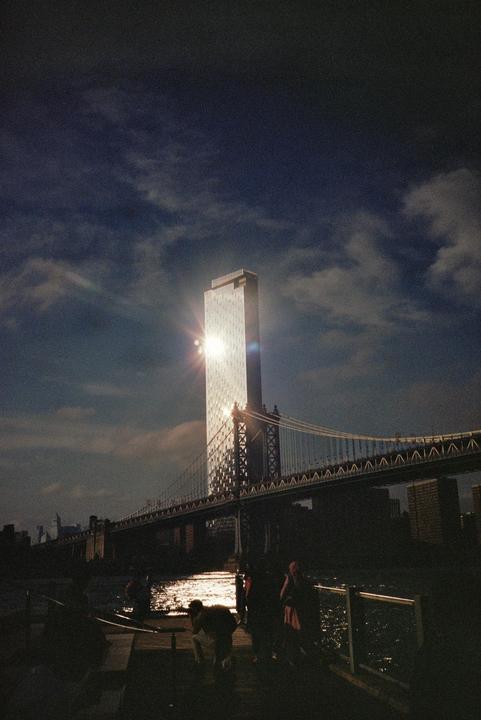 A tall building with sun glare on its facade stands next to a suspension bridge over water, with people silhouetted in the foreground.