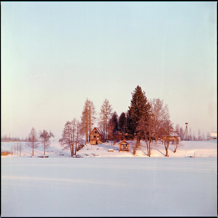 A snow-covered landscape features a frozen lake in the foreground and a cluster of houses and trees on a snowy hill in the background.
