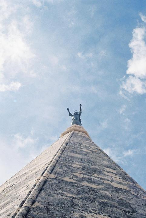 A statue with arms raised to the sky stands atop a stone pyramid against a cloudy blue sky.