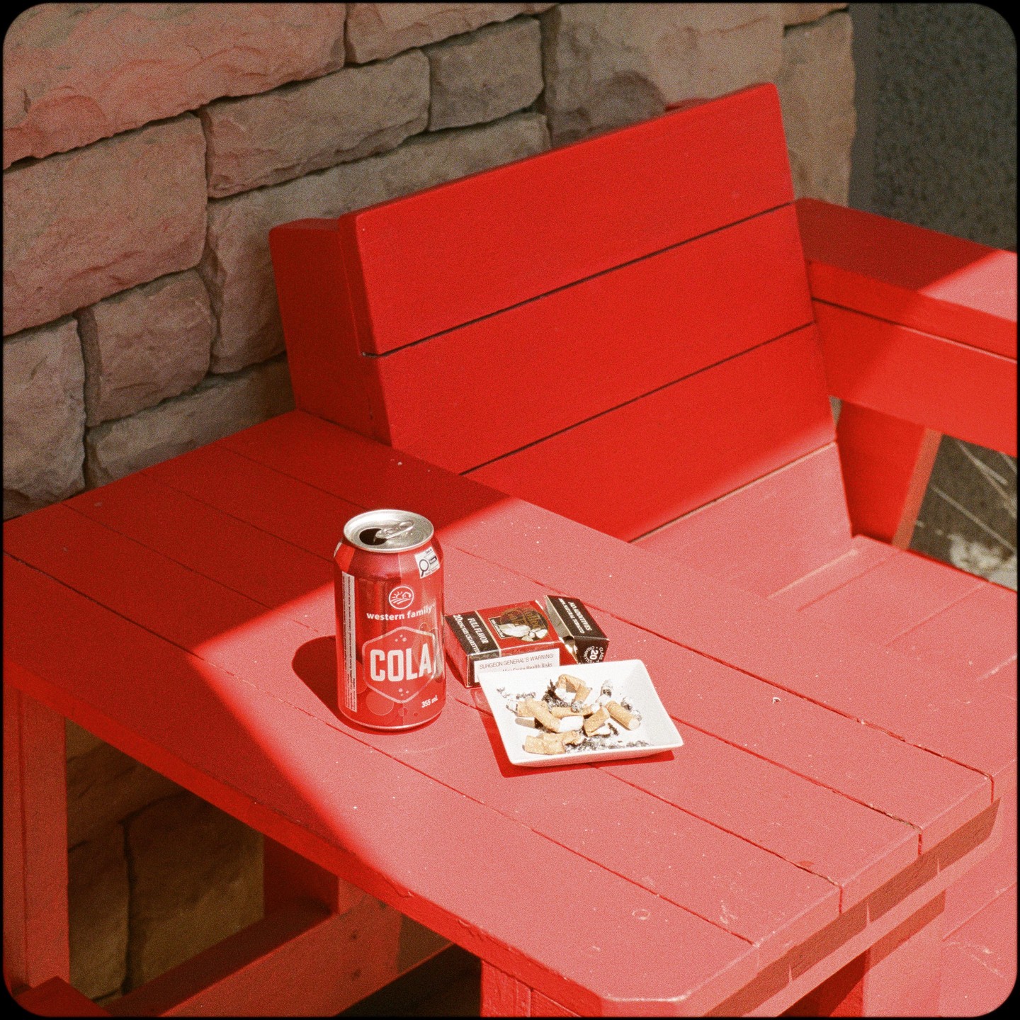 A red can of cola, a pack of cigarettes, and a plate of cigarette butts sit on a red wooden table next to a red chair.