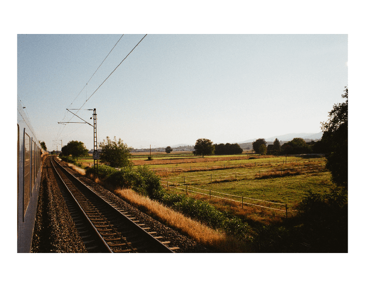 A train travels along a track next to a field with trees and hills in the distance.