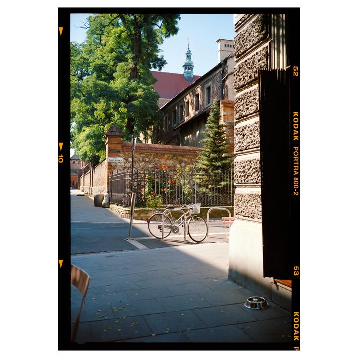 A bicycle is parked on a sunlit sidewalk next to a stone wall with a building and trees in the background.