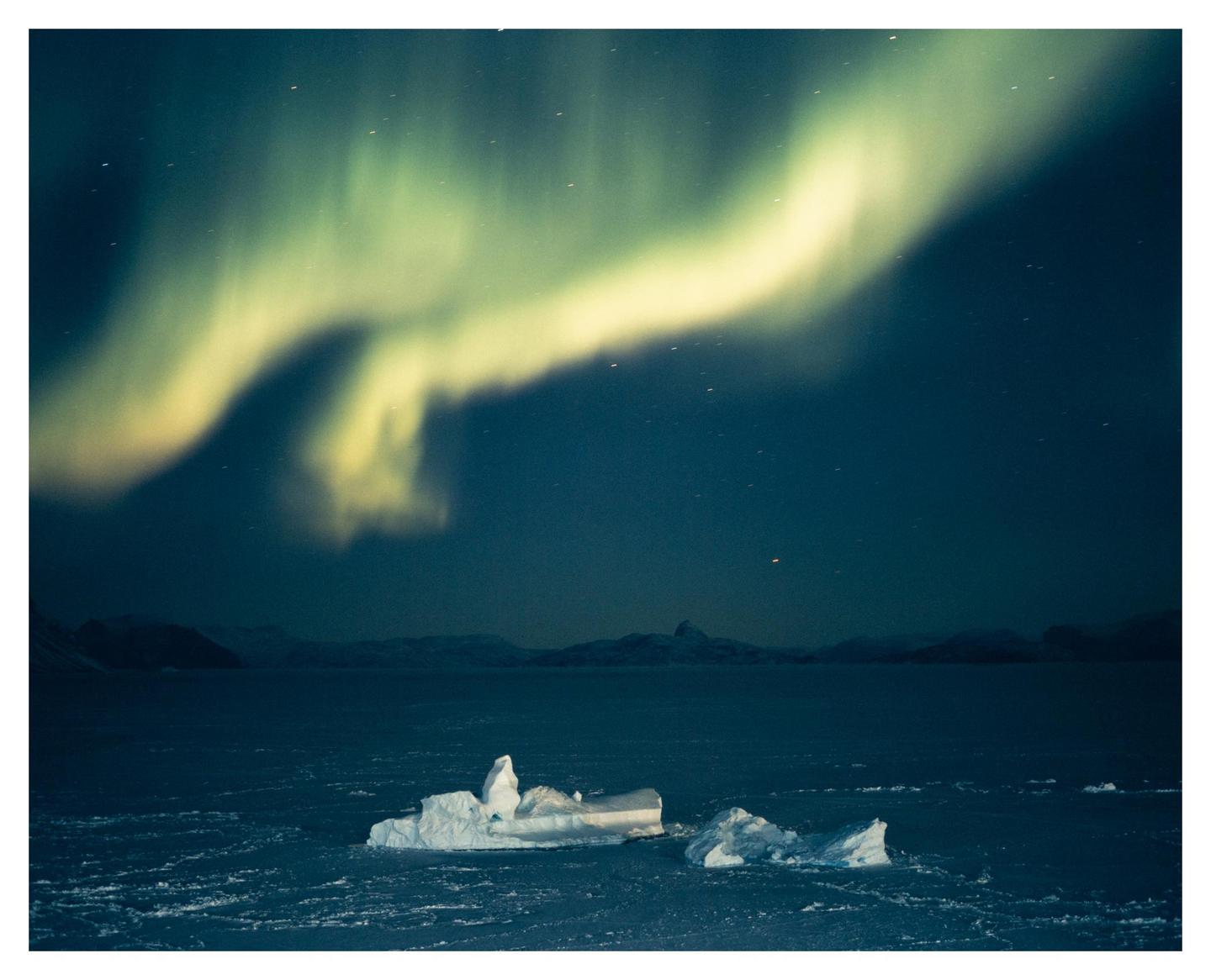 The aurora borealis glows above a dark landscape with icebergs in the water.