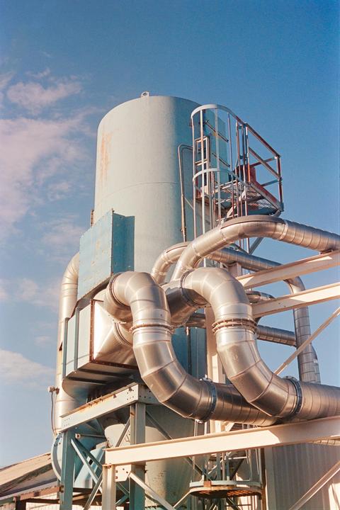A large industrial machine with pipes and a ladder is set against a blue sky with clouds.