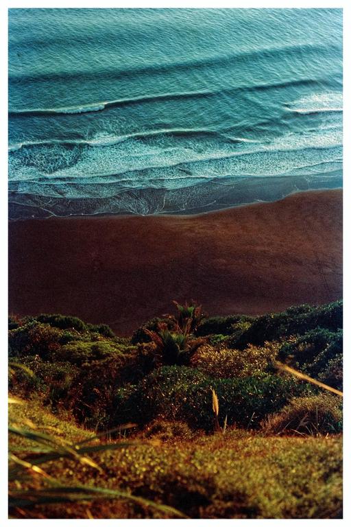 An aerial view shows waves breaking on a dark sand beach next to a lush green hillside.