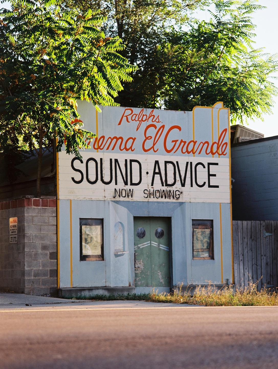 A small, light blue building with a green door and a sign that reads "Ralphs Cinema El Grande SOUND ADVICE NOW SHOWING" stands next to a road.