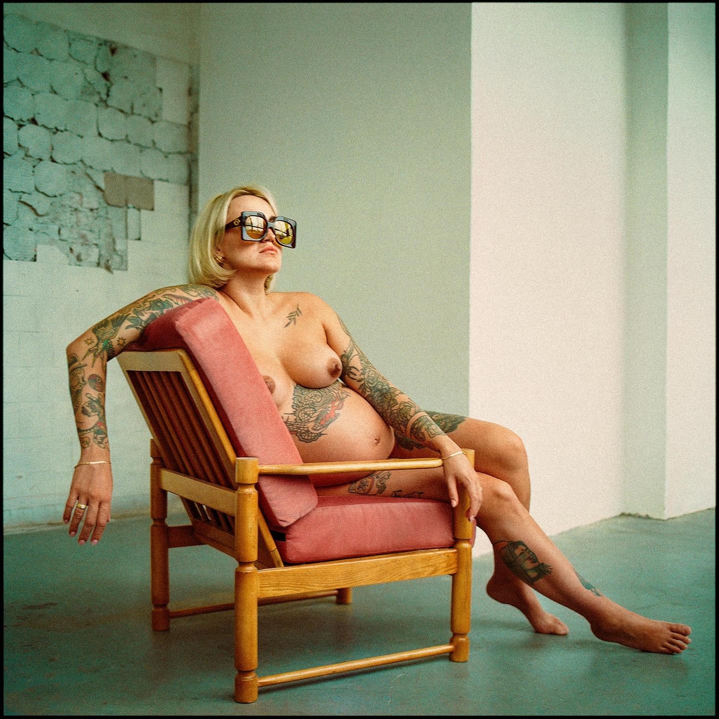 A pregnant woman with tattoos and sunglasses sits in a wooden chair with pink cushions.