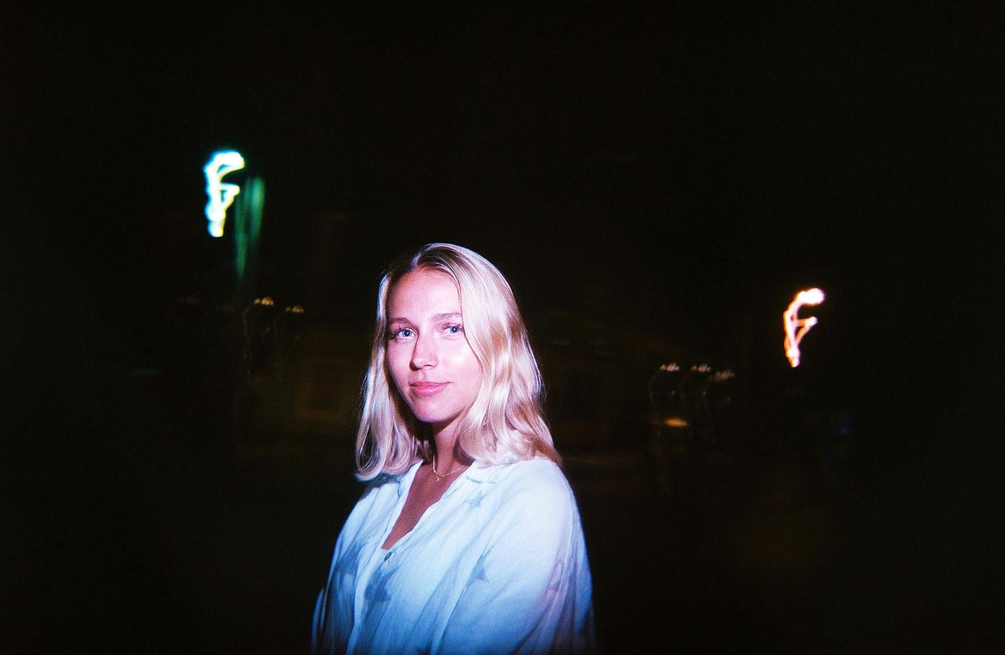 A woman with blonde hair looks at the viewer in a dark setting with blurred lights in the background.
