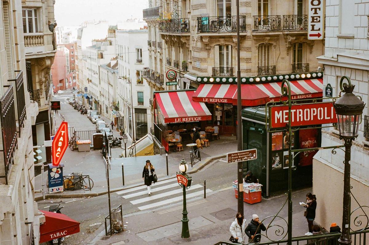 A woman crosses a street with a cafe and a metro station entrance on a hill in Paris.