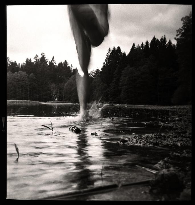A person's legs splash into a body of water with a forest in the background.