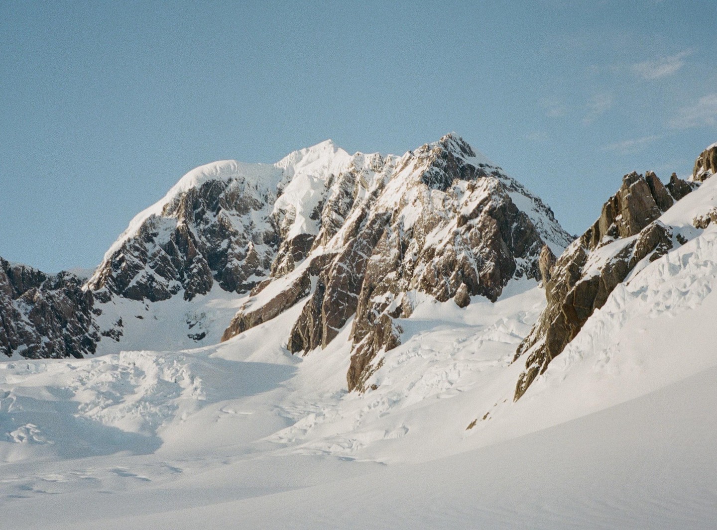 Snow-covered mountains rise under a clear blue sky, with rocky outcrops visible through the snow.