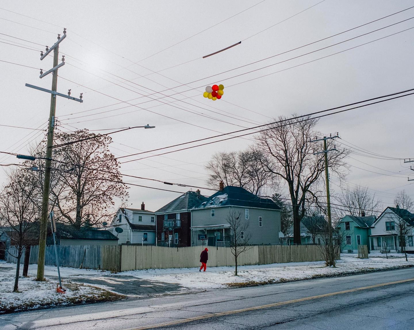 A person in a red coat walks along a snowy street with houses and bare trees, while balloons are tangled in overhead power lines.