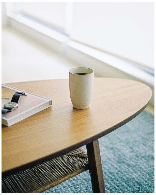 A cup of coffee sits on a wooden table next to a book and a watch.
