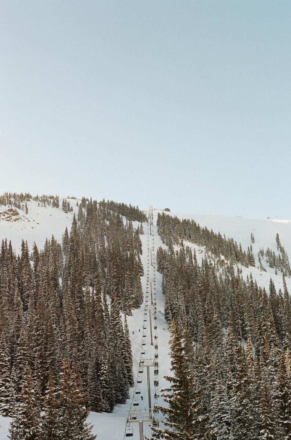 A ski lift ascends a snow-covered mountain surrounded by pine trees under a clear sky.