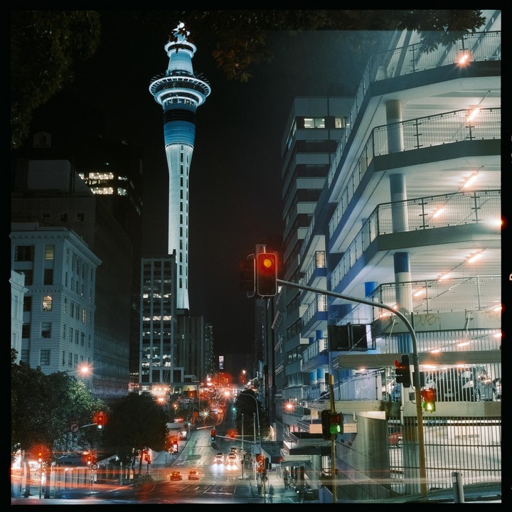 A tall, illuminated tower stands above a city street with traffic lights and buildings at night.