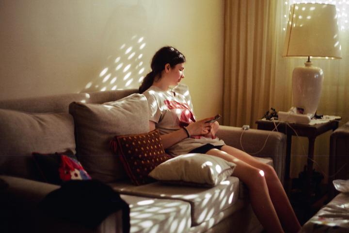 A young woman sits on a sofa in a living room, illuminated by dappled sunlight.