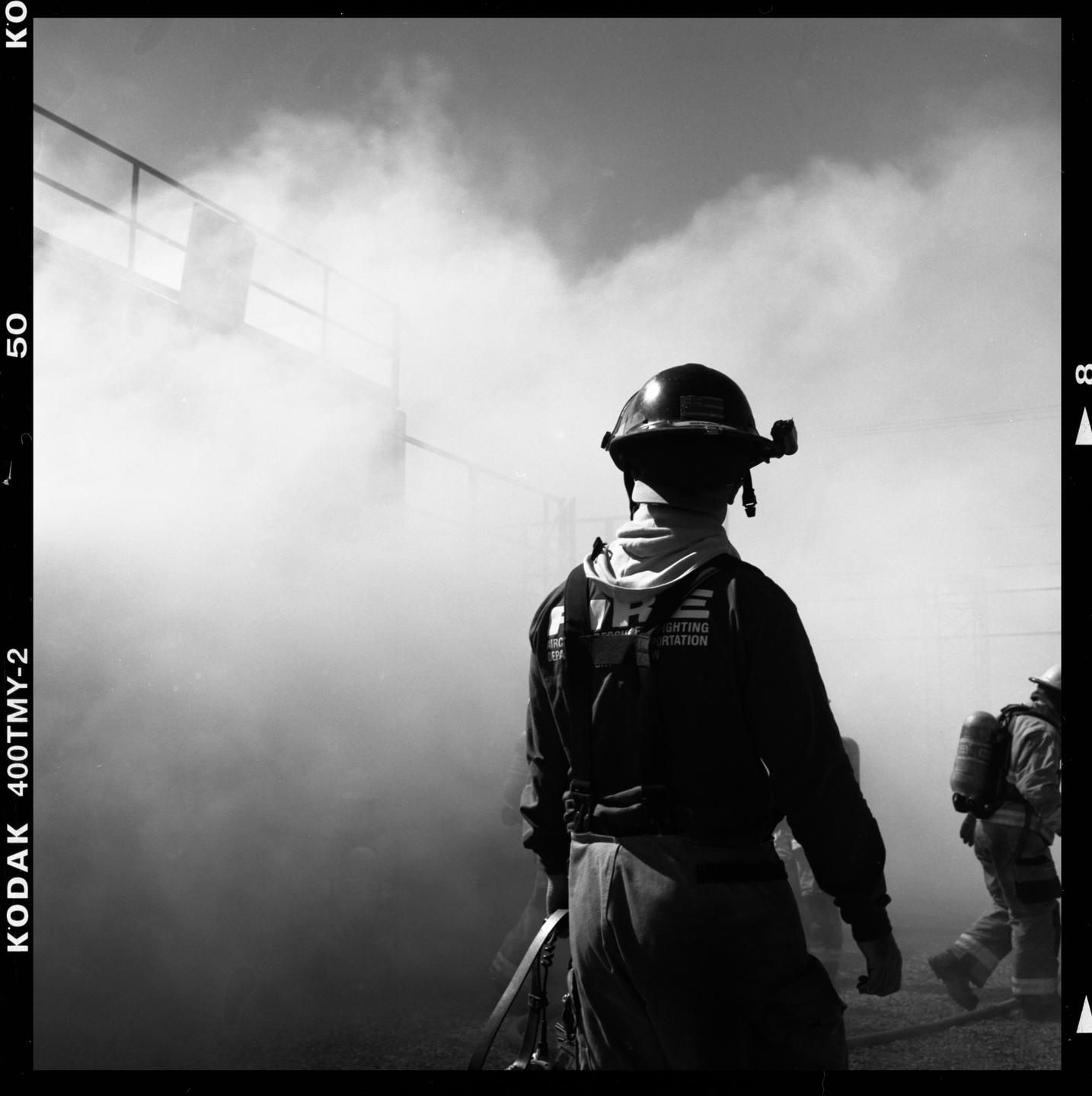 A firefighter wearing a helmet and uniform stands with their back to the viewer, surrounded by smoke.
