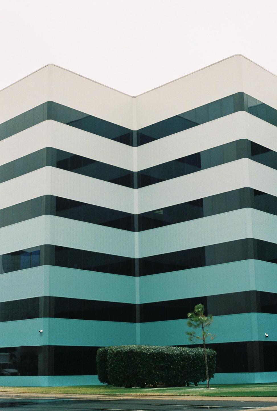 A modern building with horizontal stripes of windows and light blue walls stands behind a manicured hedge and a small tree.