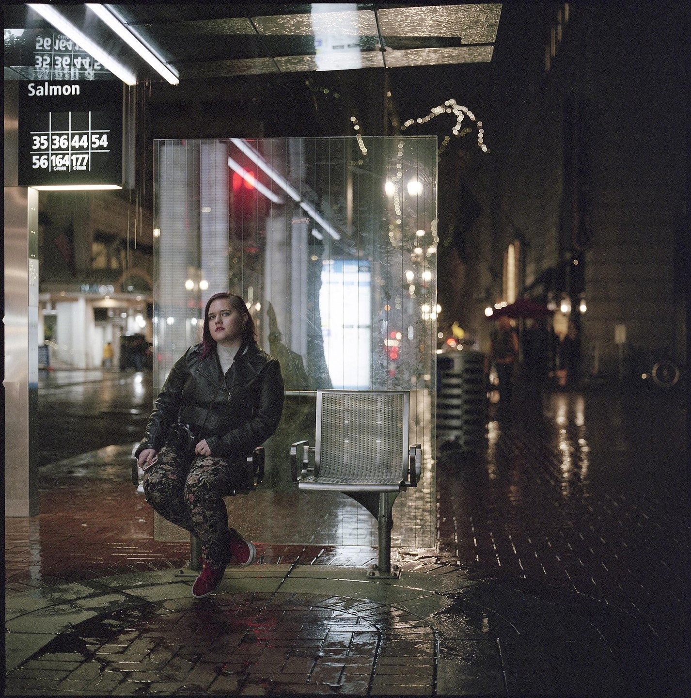 A woman sits at a bus stop on a wet city street at night.