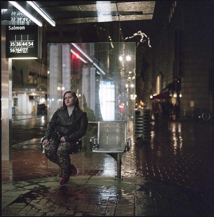 A woman sits at a bus stop on a wet city street at night.