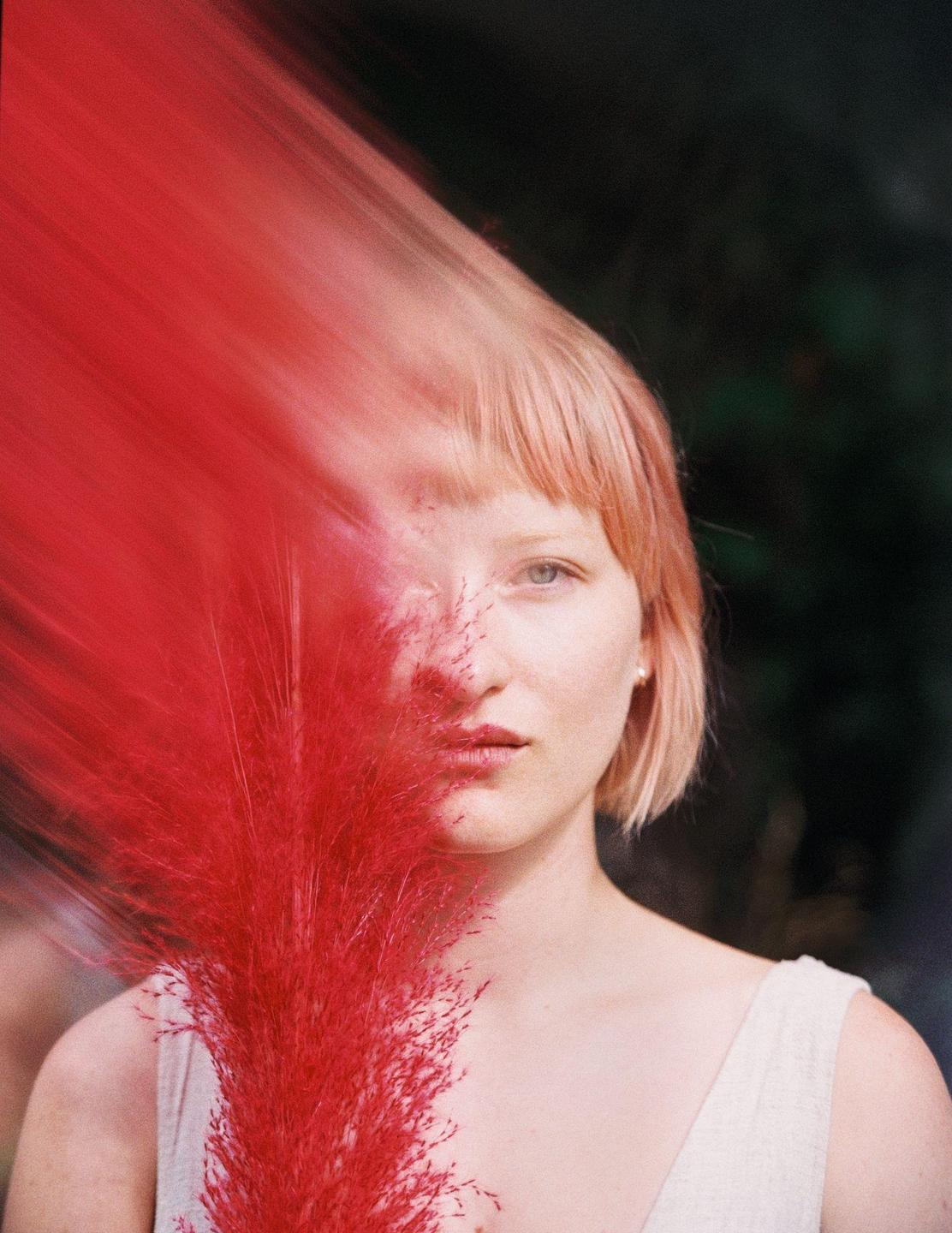 A woman with short blonde hair looks directly at the viewer, with a bright red plant partially obscuring her face.