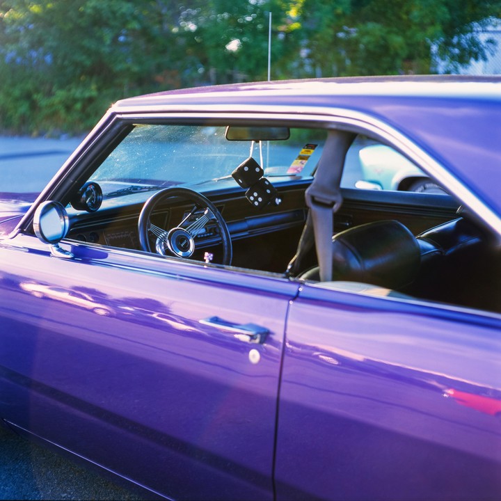 A purple car with fuzzy dice hanging from the rearview mirror is parked on a street.