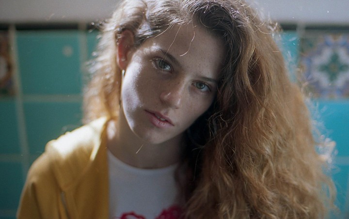 A woman with long, wavy hair and freckles looks directly at the viewer, with a blue tiled wall behind her.