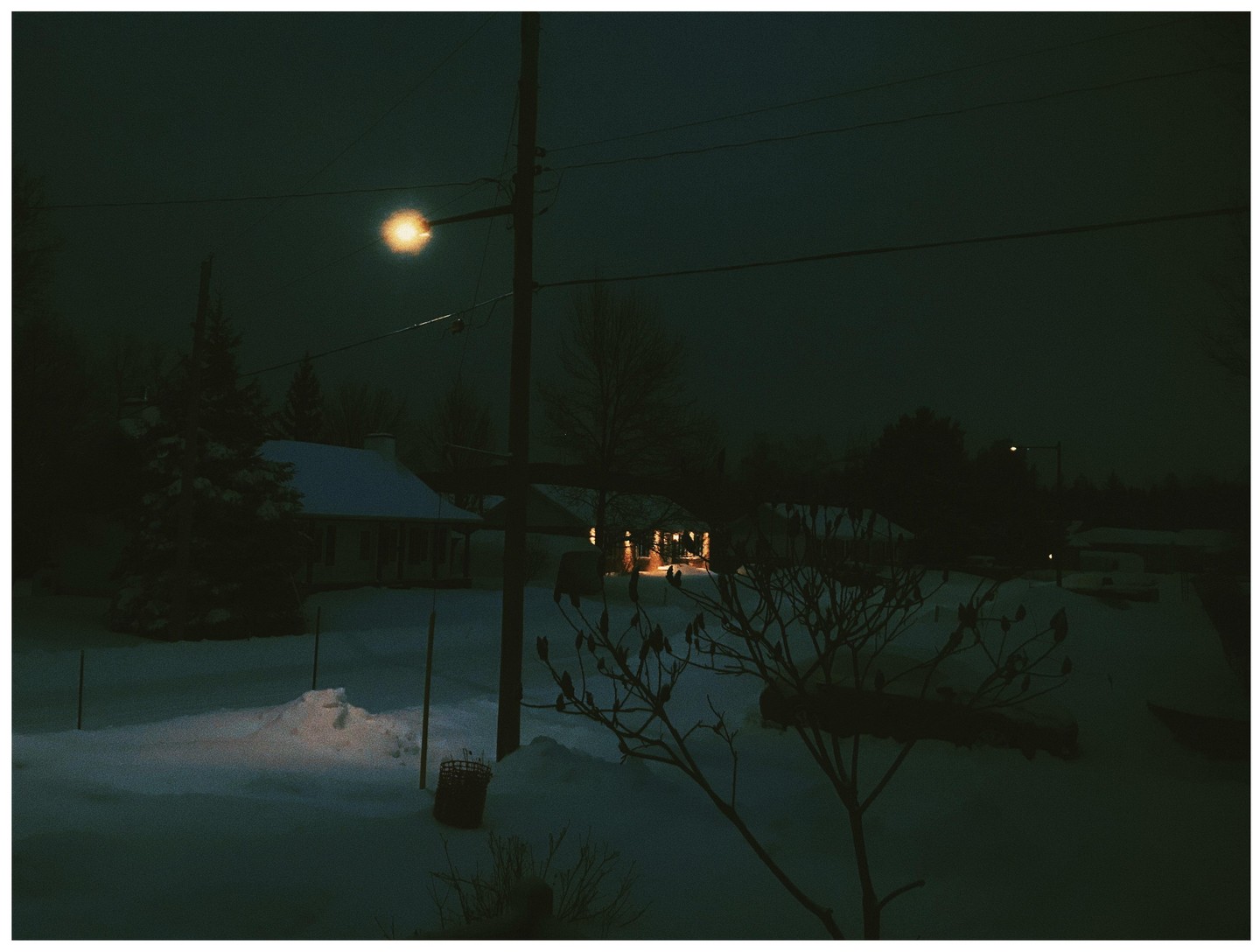 A snow-covered neighborhood is illuminated by a street light and house lights at night.