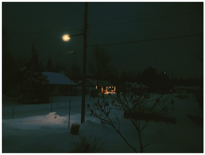 A snow-covered neighborhood is illuminated by a street light and house lights at night.