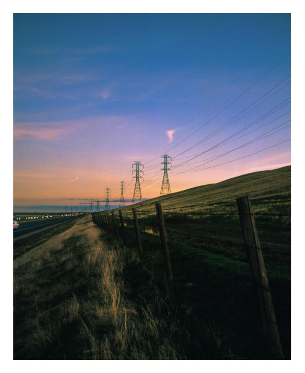A row of power line towers stands on a grassy hill next to a highway at dusk.