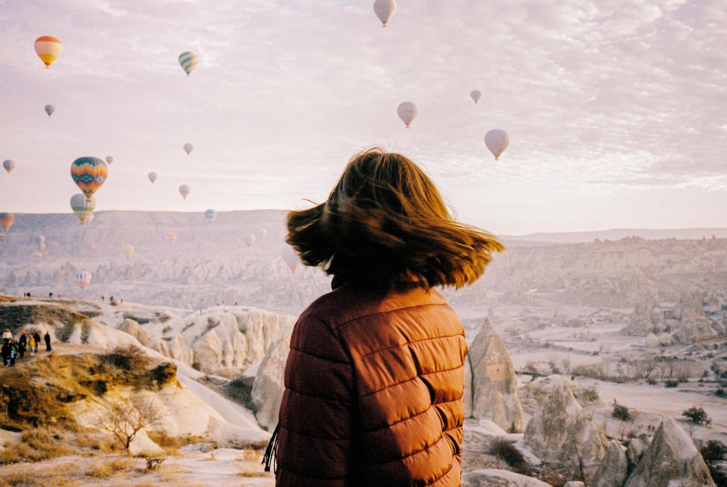 A woman with windblown hair stands on a hill overlooking a valley filled with hot air balloons and rock formations.