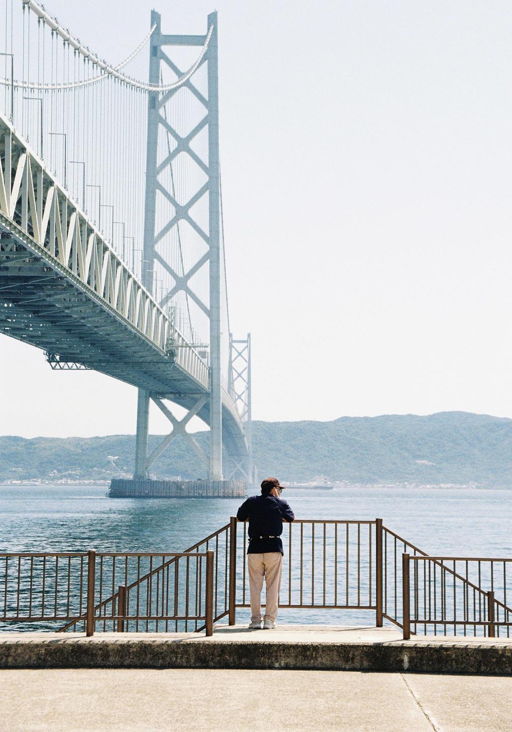 A man stands on a concrete platform looking out at a large suspension bridge over water with mountains in the distance.